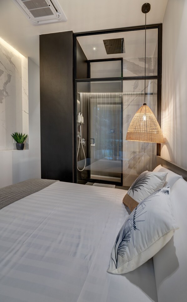 A modern bedroom with a large bed, a glass shower enclosure, a hanging light fixture, and a small potted plant.