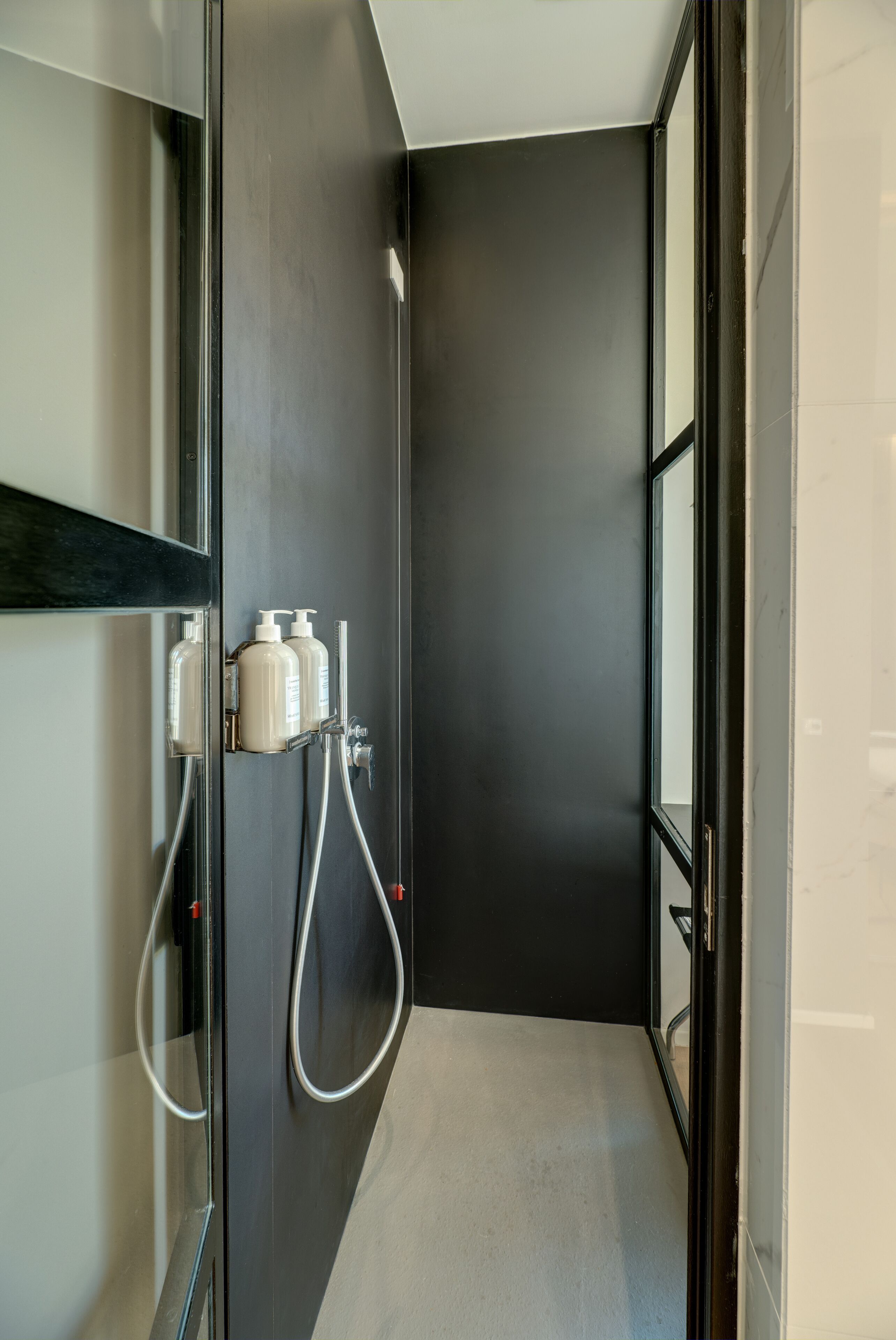 A modern bathroom with a dark wall, a glass shower enclosure, and two white bottles on the wall.