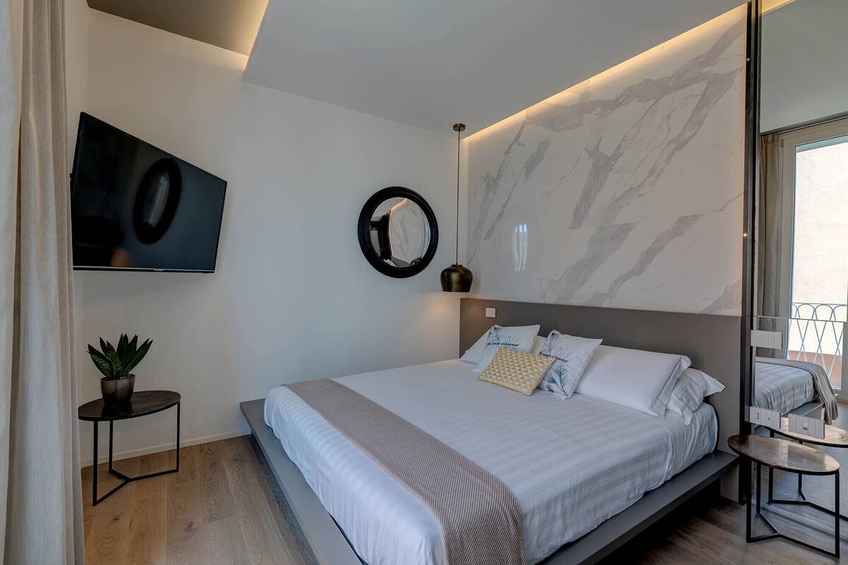A modern bedroom with a large bed, a TV mounted on the wall, a round mirror, and a small table with a plant.
