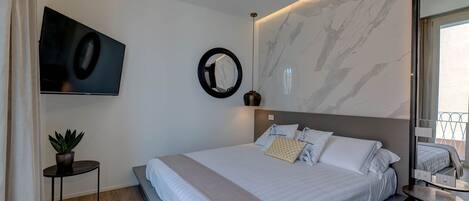 A modern bedroom with a large bed, a TV mounted on the wall, a round mirror, and a small table with a plant.