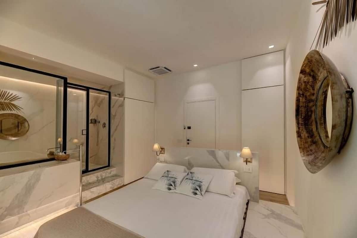 A modern bedroom with a large bed, a walk-in closet, a bathroom with a glass shower, and a large mirror on the wall.