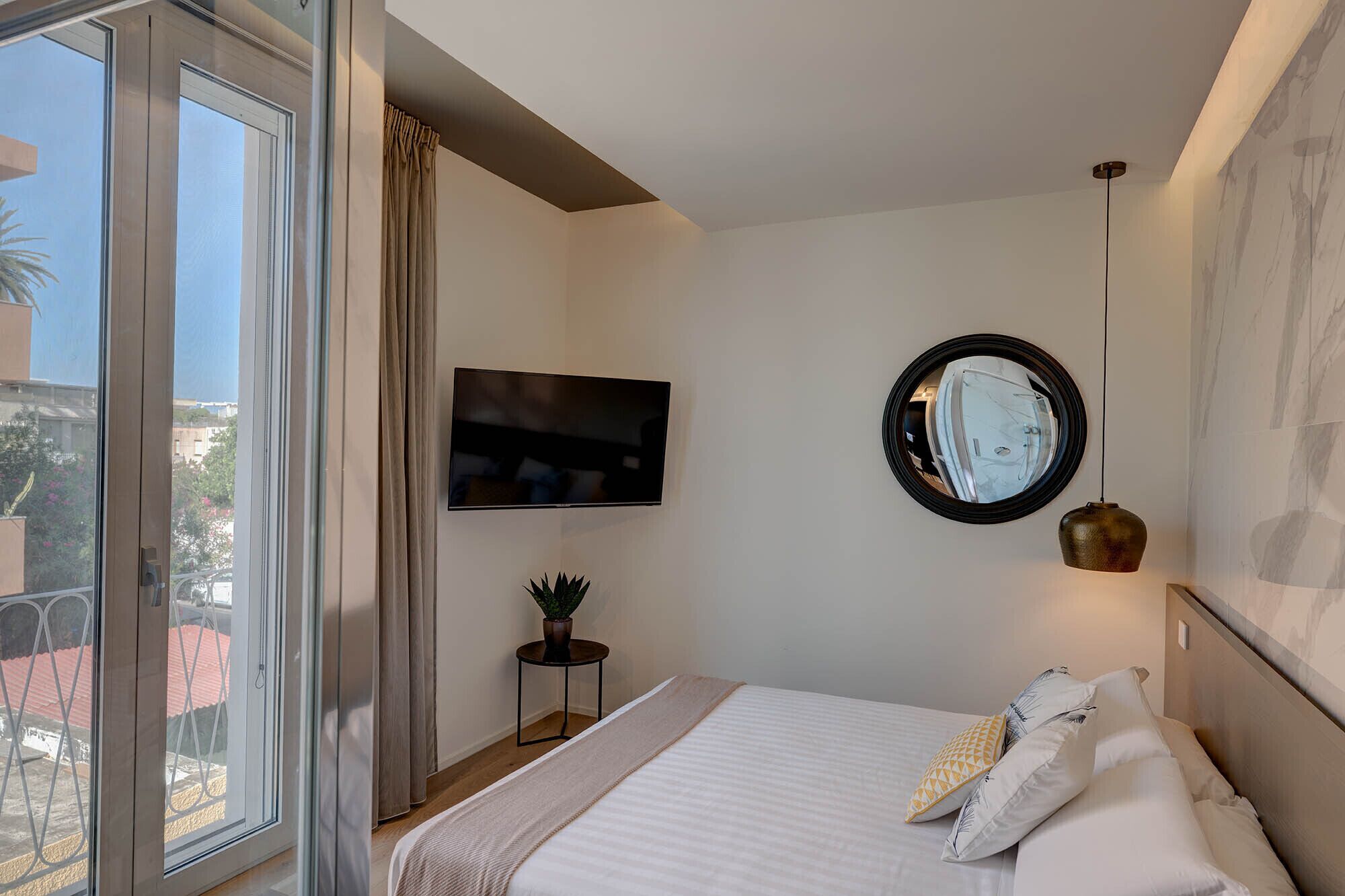 A hotel room with a bed, a TV, a round mirror, and a window with curtains.