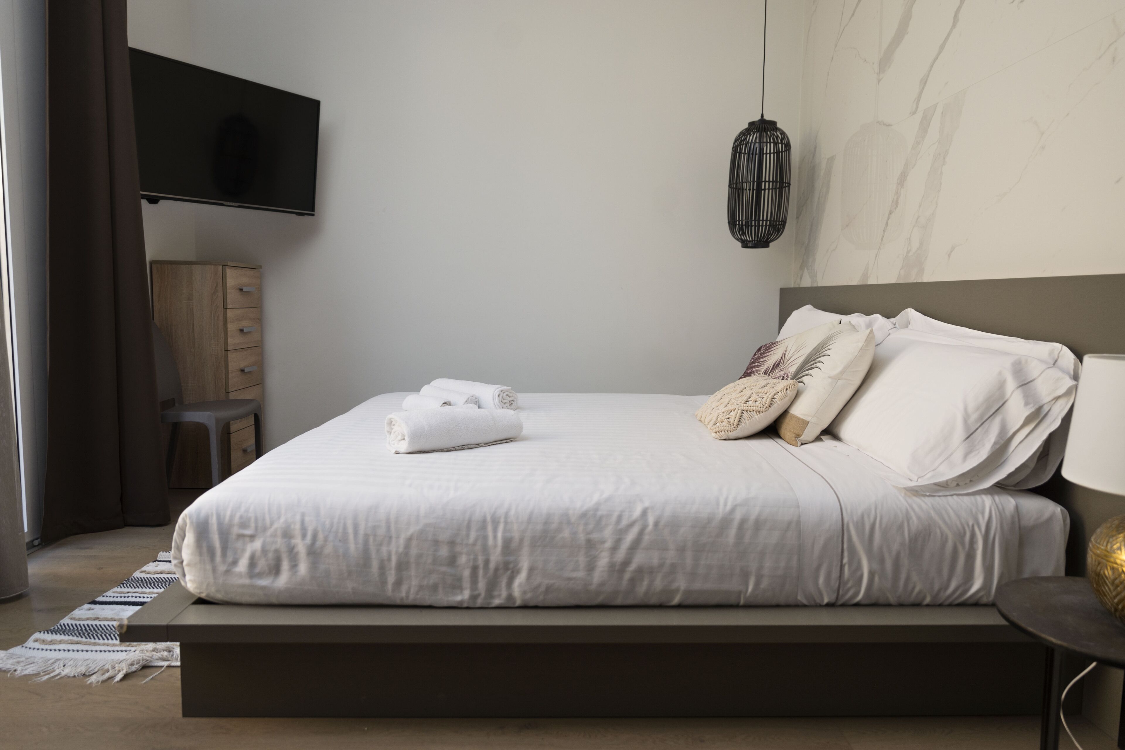 A modern bedroom with a large bed, a flat-screen TV mounted on the wall, a bedside table, and a small wooden cabinet.