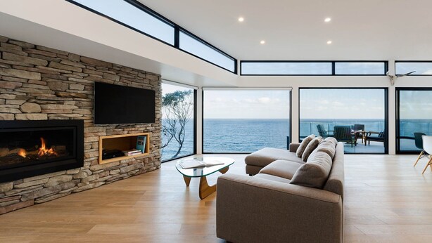 A modern living room with a stone fireplace, a flat-screen TV, and a large window overlooking the sea. - Sea Ranch | Popular Coastal Retreat with Ocean View Bath (Wye River)