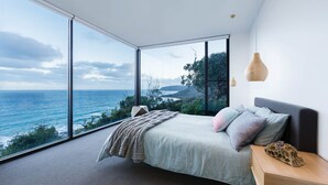 A bedroom with a large window overlooking the ocean, a bed with pillows, a wooden nightstand, and a hanging light fixture. - Sea Ranch | Popular Coastal Retreat with Ocean View Bath (Wye River)