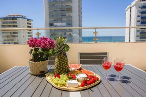 A table with a fruit platter, a pineapple, a potted plant, and two glasses of red drink on a balcony overlooking a beach.
