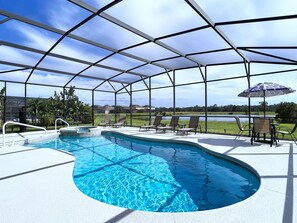 Piscina techada con sillones de descanso y una mesa, rodeada por una cerca de metal, con vista a casas y un lago al fondo.