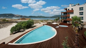 A swimming pool with a wooden deck, a beach view, and a modern building in the background.