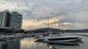 A marina with multiple yachts docked, a modern building with balconies, and a mountainous backdrop under a cloudy sky.