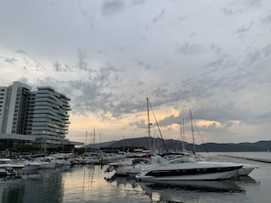 Uma marina com vários iates atracados, um edifício moderno com varandas e um cenário montanhoso sob um céu nublado.