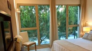 A bedroom with a large window, a bed, a chair, a lamp, and a vase with flowers.