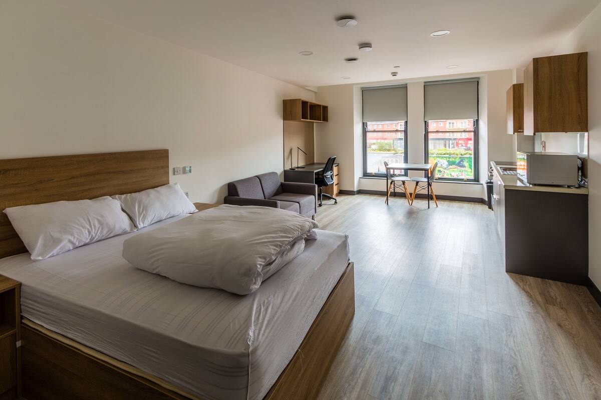 Ein modernes Hotelzimmer mit einem großen Bett, einem Schreibtisch, einem Sessel und Blick auf die Stadt durch das Fenster.