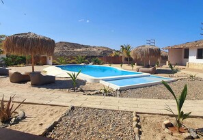 A pool area with a clear blue pool, surrounded by thatched umbrellas, and a pebble pathway leading to a building with a white exterior. - Oasis de Vichayito (Los Organos)