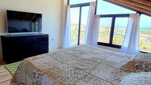 Quarto com cama grande, televisão em cômoda e vista para a paisagem através de grandes janelas.