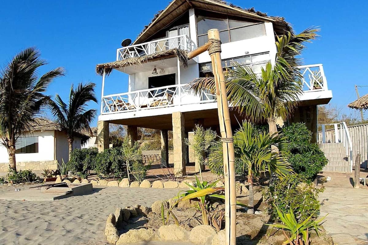 A two-story house with a balcony, surrounded by palm trees and a sandy ground.