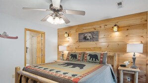 A bedroom with a wooden headboard, a bed with a bear-themed bedspread, two bedside lamps, a ceiling fan, and a wooden door. - River Bear-Riverfront in Pigeon Forge (Pigeon Forge)