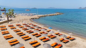 A beach with rows of orange sun loungers and umbrellas, a rocky breakwater, and a sailboat in the distance. - Asterina Hotel (Bodrum)