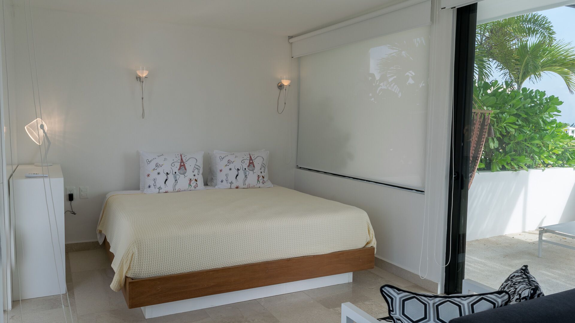 Une chambre à coucher avec un lit, deux coussins décoratifs, des lampes de chevet, une armoire blanche et une vue sur la verdure à l’extérieur.