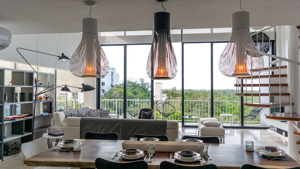 A modern dining area with a wooden table set for four, surrounded by a sofa and chairs, with large windows offering a view of greenery and buildings. - Siaan Betine 4br Deluxe PDC  (Playa del Carmen)