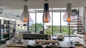 A modern dining area with a wooden table set for four, surrounded by a sofa and chairs, with large windows offering a view of greenery and buildings. - Siaan Betine 4br Deluxe PDC  (Playa del Carmen)