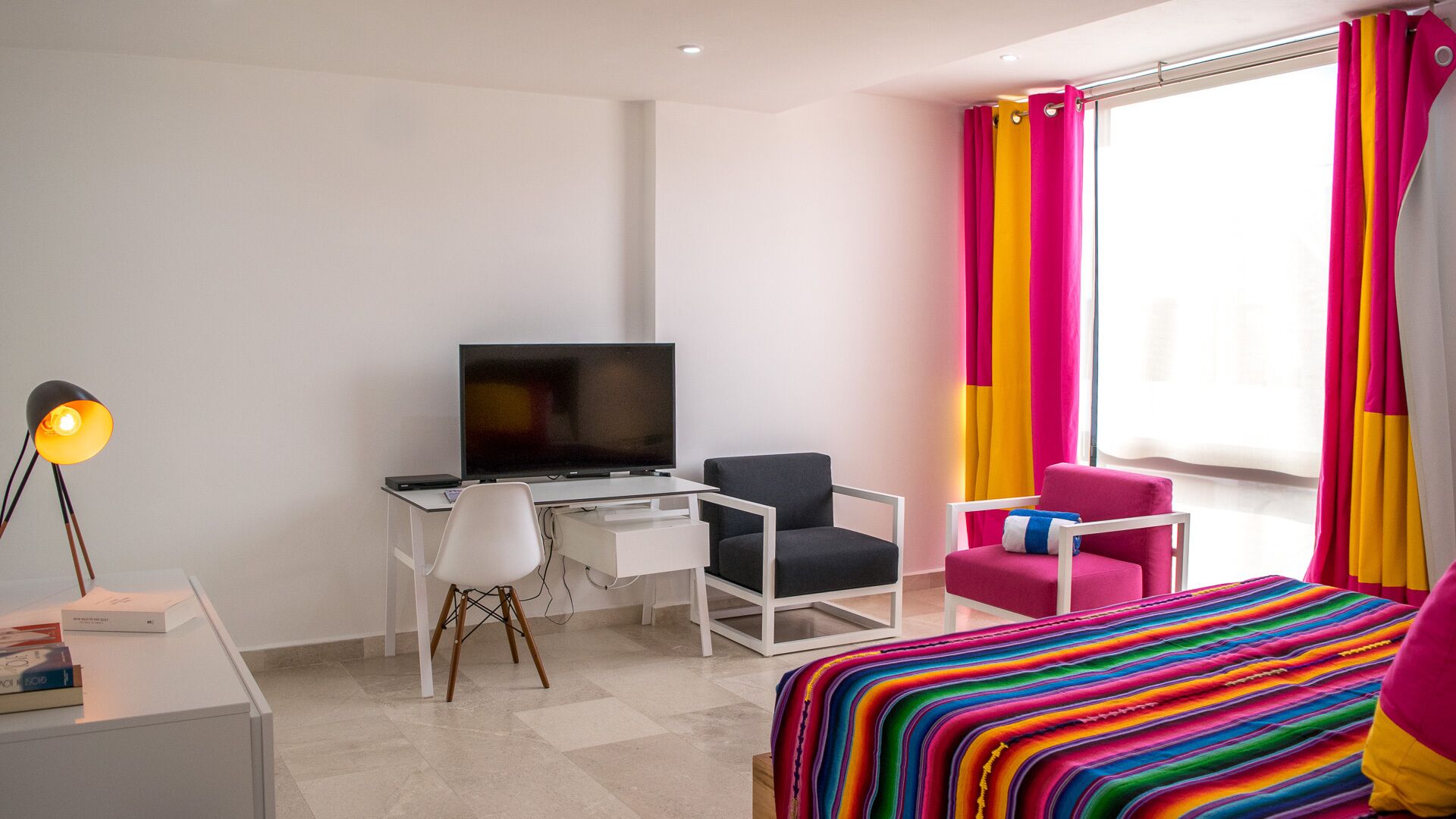 Una habitación de hotel moderna con una cama, un escritorio con televisor, una silla y una manta a rayas colorida.