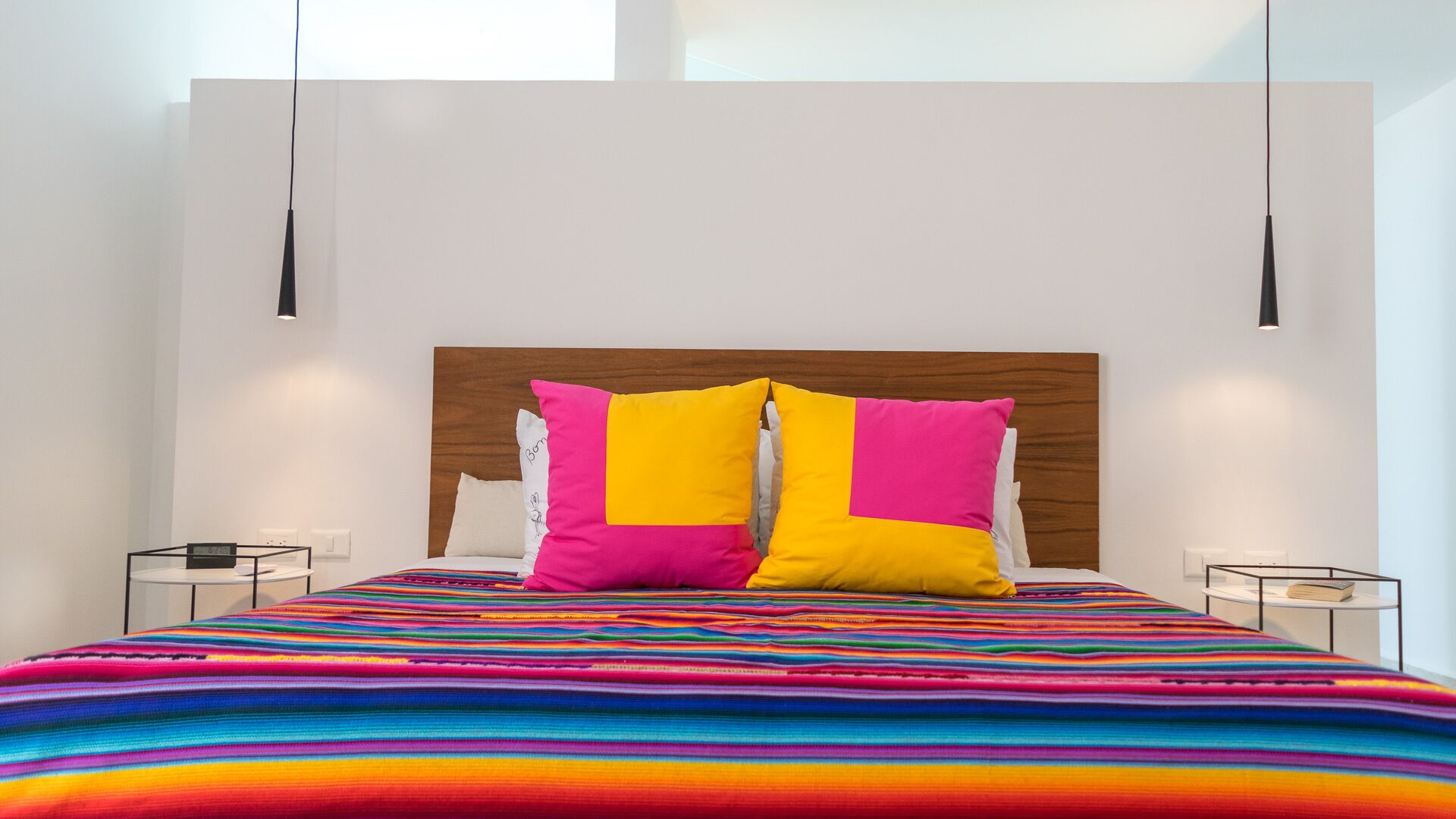 A modern bedroom with a wooden headboard, a colorful striped bedspread, and two bedside tables with lamps.