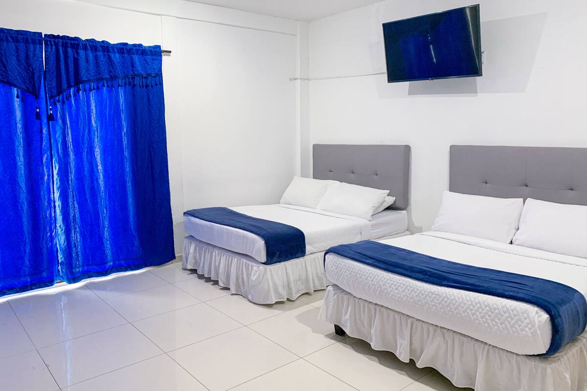 Habitación de hotel con dos camas, un televisor de pantalla plana montado en la pared y un gran ventanal con cortinas azules.