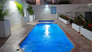 A swimming pool with a tiled deck, surrounded by white planters with red flowers, and a cityscape in the background. - Hotel Rodadero Inn By GEH Suites (Santa Marta)