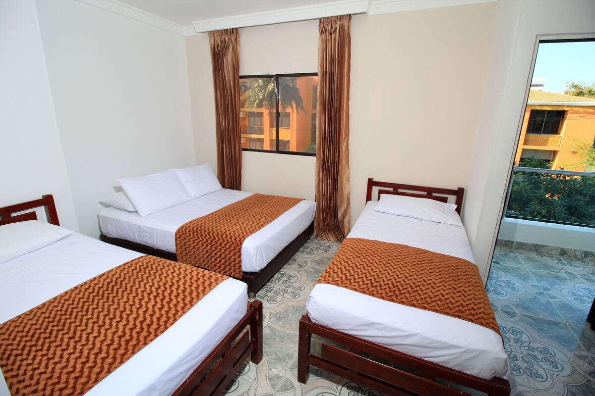 A hotel room with three single beds, each with a patterned bedspread, a window with curtains, and a view of a building outside.