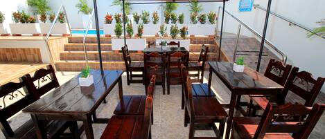 An outdoor patio with wooden tables and chairs, surrounded by potted plants and a swimming pool in the background.