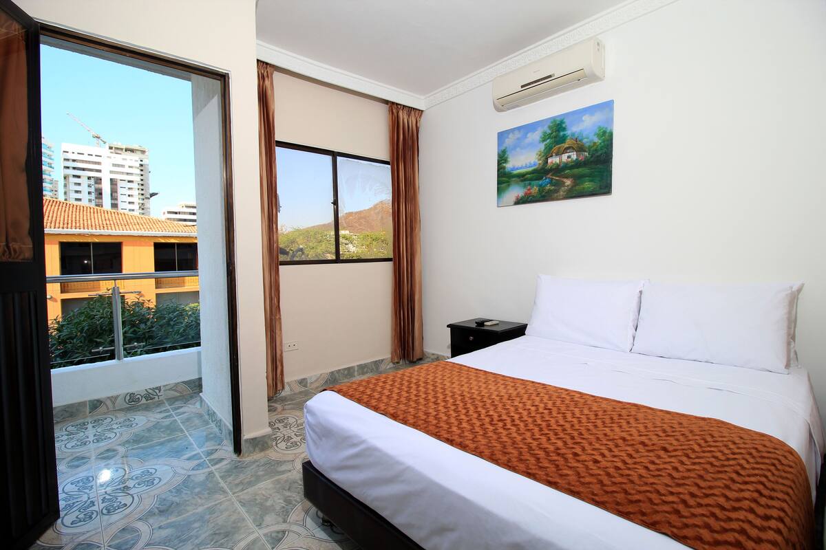 A hotel room with a large bed, a painting on the wall, a window with curtains, and an air conditioning unit.
