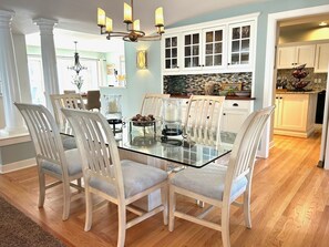A dining area with a glass-top table surrounded by white chairs, white cabinets, and a chandelier.