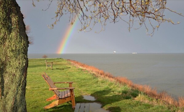 A wooden chair by a grassy lakeside with a rainbow in the background.