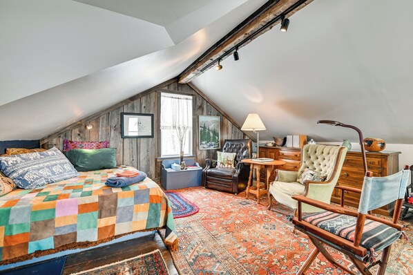 A cozy attic bedroom with a sloped ceiling, a large bed with a colorful quilt, a leather armchair, and a wooden dresser. - Cozy Catskills Vacation Rental with Deck! (Fleischmanns)
