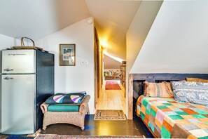 A compact room with a bed, a wicker chair, a small fridge, and a wooden door leading to another room. - Cozy Catskills Vacation Rental with Deck! (Fleischmanns)