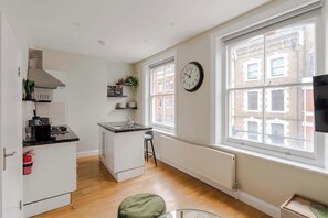 A compact kitchen with a central island, a large window with a view of buildings, a wall clock, and a radiator.