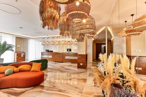 A modern lobby with a reception desk, a plush orange sofa, and a decorative hanging light fixture. - Hotel Tq Plaza (Budva)