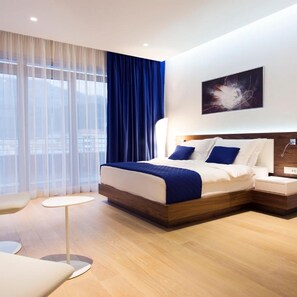 A modern hotel room with a large bed, a bedside table, a nightstand, a chair, and a view of the city through large windows. - Hotel Tq Plaza (Budva)