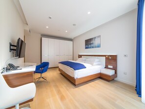 A modern hotel room with a wooden bed, a desk with a computer monitor, a chair, and a wardrobe.