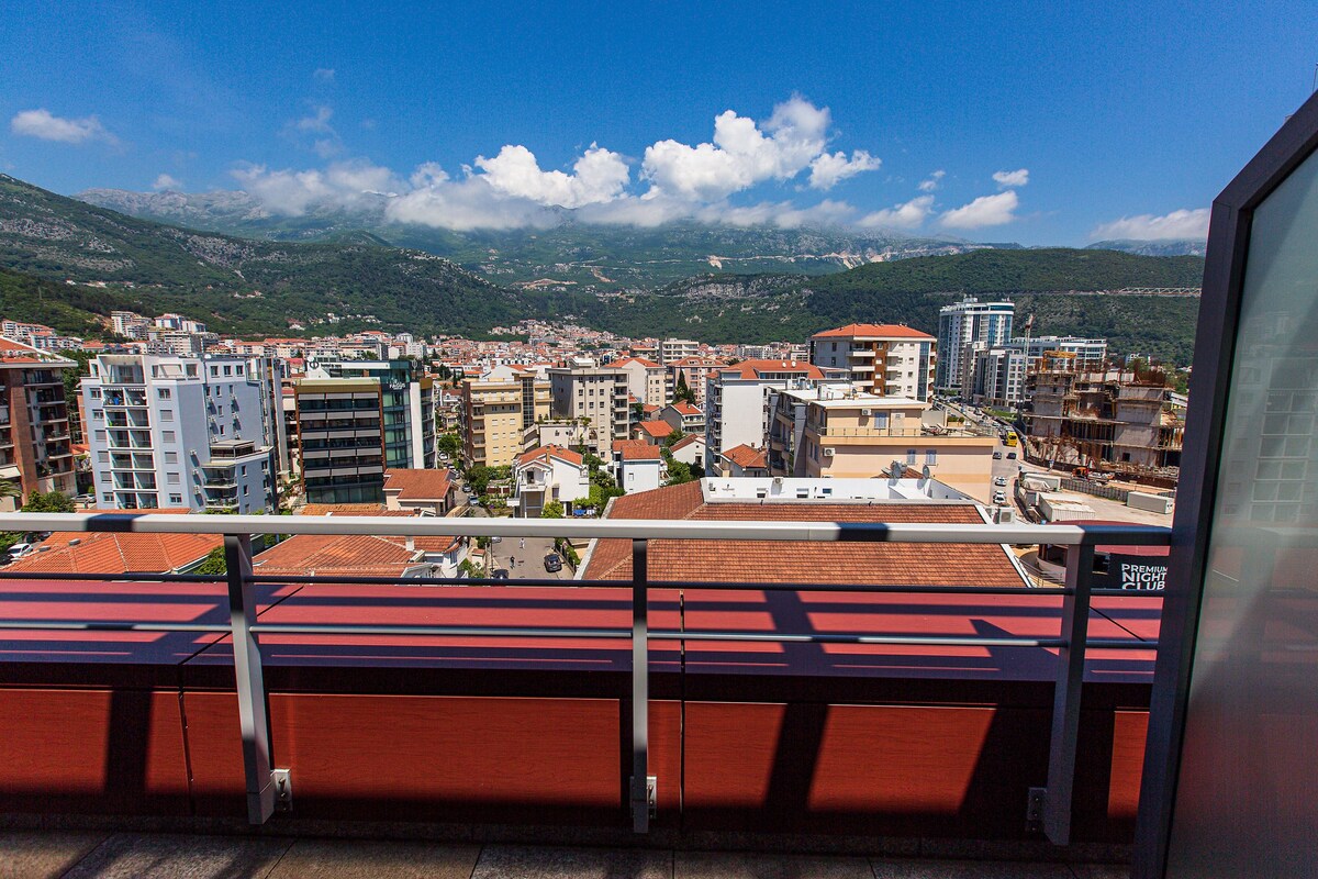A cityscape with a view of mountains in the background, buildings with red roofs, and a balcony with a metal railing.