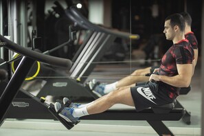 Un homme utilise un appareil de rame dans une salle de sport, avec d’autres équipements d’exercice en arrière-plan.