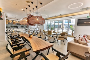 A modern dining area with long wooden tables, woven pendant lights, and a view of the city through large windows.