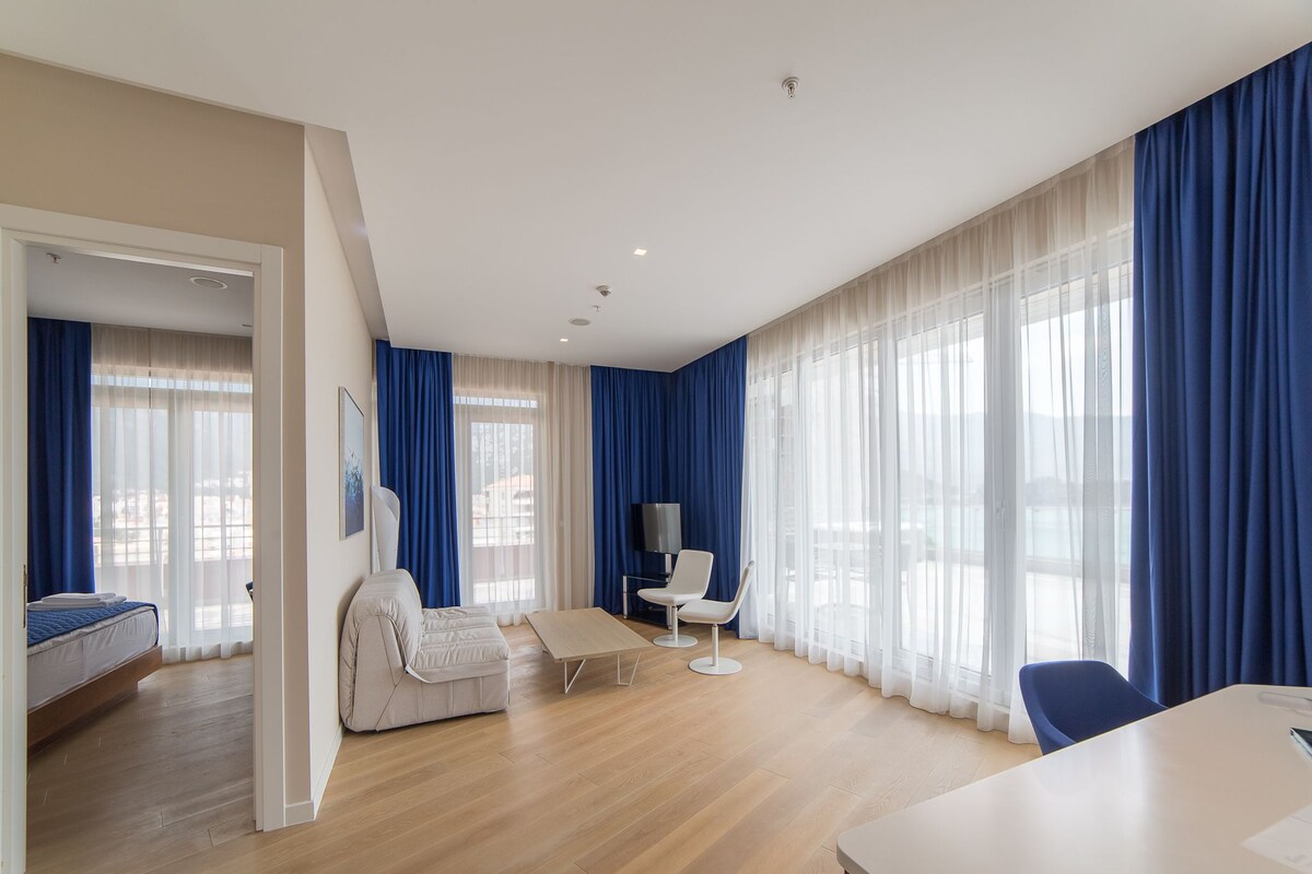 A modern hotel room with a bed, a sofa, a dining table, and a TV.