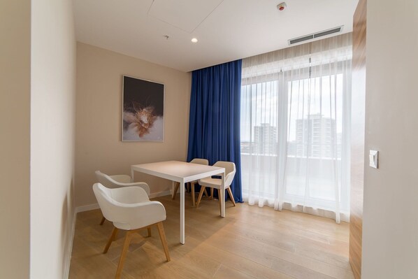 A modern, well-lit room with a dining table and chairs, large windows with sheer curtains, and a city view.