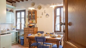 Un rincón comedor acogedor con una mesa de madera preparada para cuatro personas, rodeada de sillas azules. En el fondo se observa un aparador de madera con puertas de vidrio, y una ventana con cortinas deja entrar luz natural.