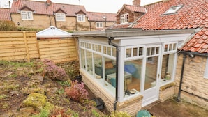 A conservatory with stone walls and a tiled roof, surrounded by a garden with various plants and a wooden fence. - Riseholme (Scarborough)