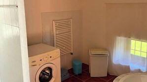 A laundry room with a washing machine, a dryer, a trash bin, and a window with blinds.