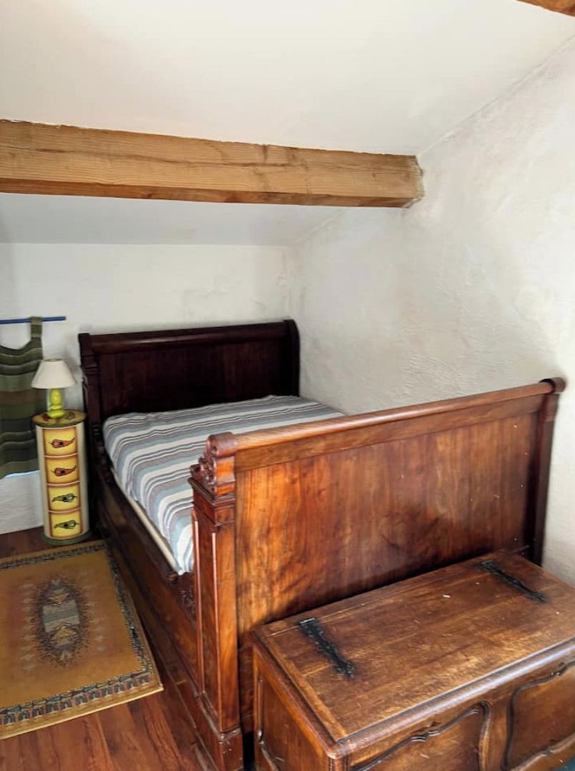 Una habitación pequeña y estrecha con una cama de madera, una mesita de noche, una lámpara y un cofre pequeño de madera.