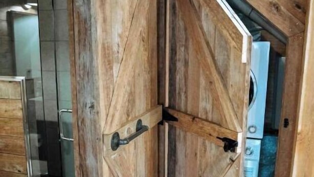 Une porte en bois rustique, dotée d’une poignée en métal, donne accès à une salle de bain avec une douche en verre et un lavabo blanc.