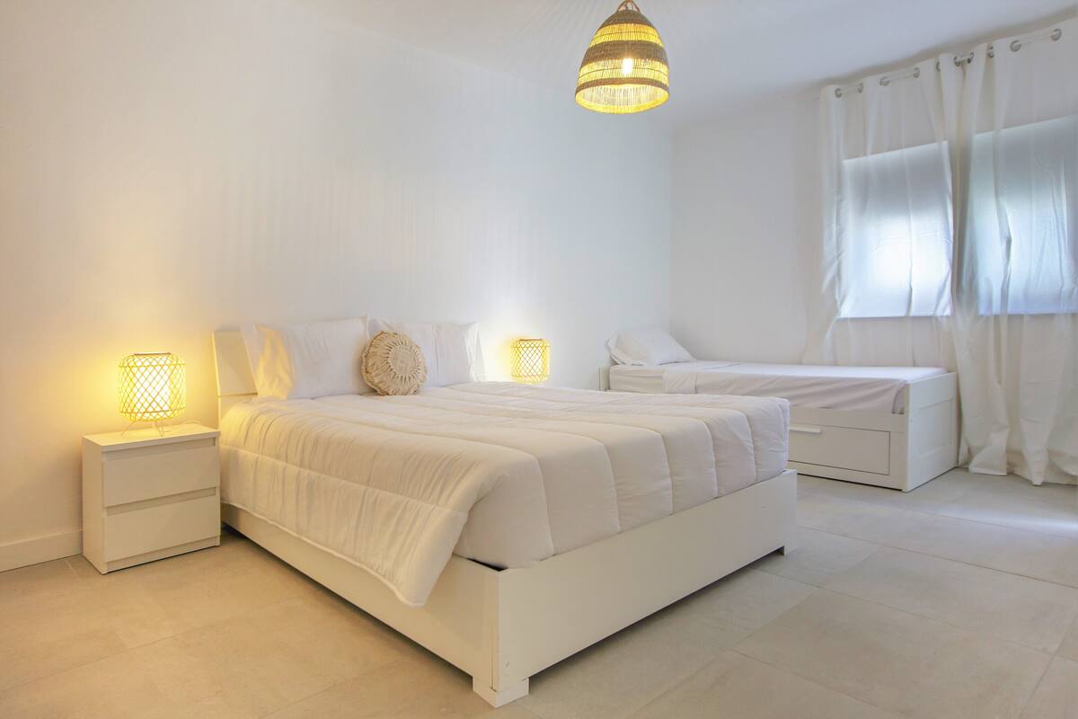 A modern bedroom with a large bed, bedside tables, a nightstand, a bedside lamp, a hanging light fixture, and a window with curtains.